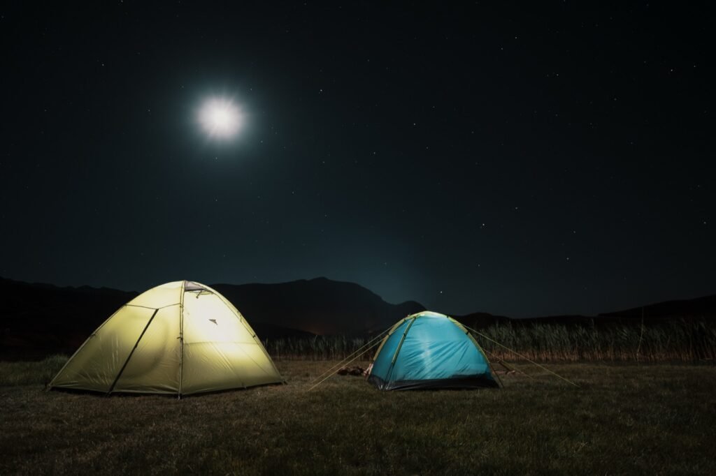 Two lit dome tents, one yellow and one blue, on a grassy meadow at night under a bright moon and stars