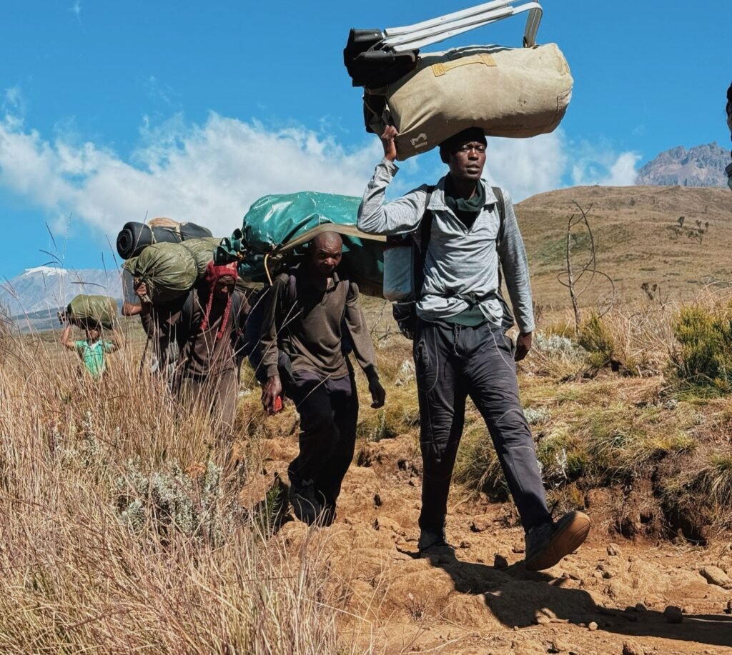 Porters carrying large duffel bags and loads on their heads and shoulders along a dry moorland trail on Kilimanjaro, with Mawenzi peak in the distance