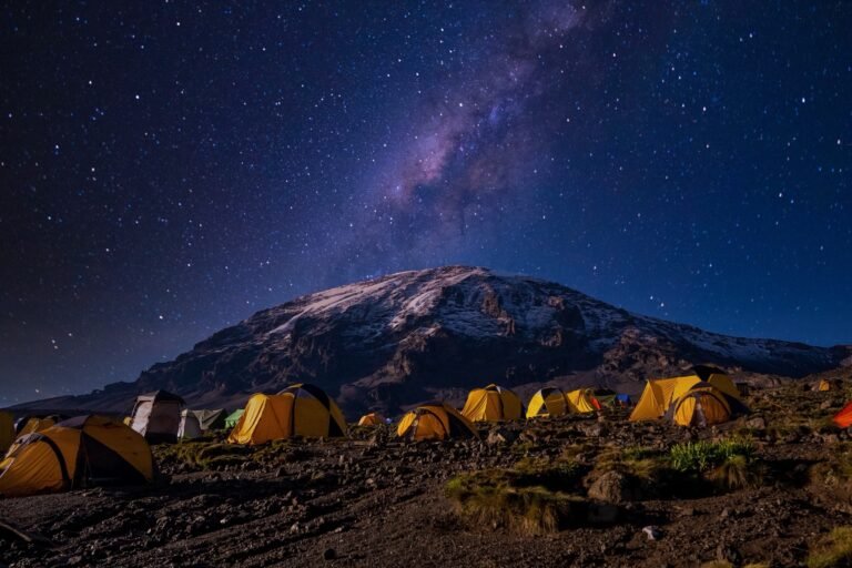 Yellow expedition tents at a rocky high camp beneath the snow-dusted summit of Kibo, with the Milky Way and a star-filled night sky above