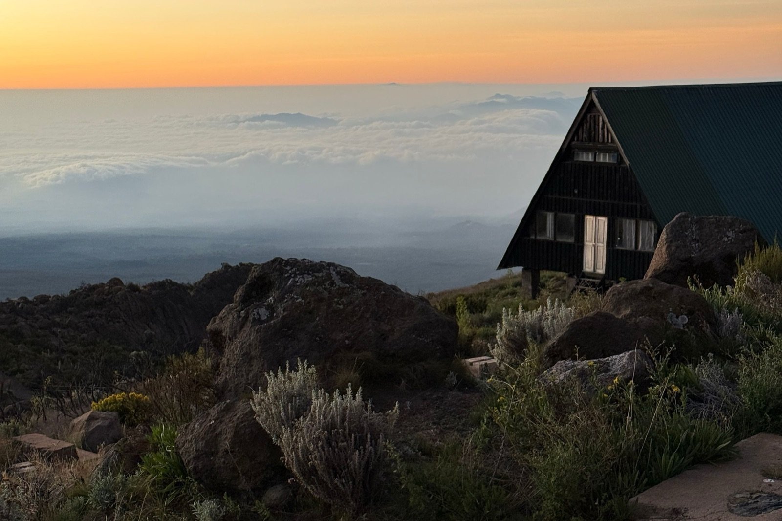 Wooden A-frame mountain hut on Kilimanjaro at sunset, above a sea of clouds, with rocks, heather and a stone path in the foreground