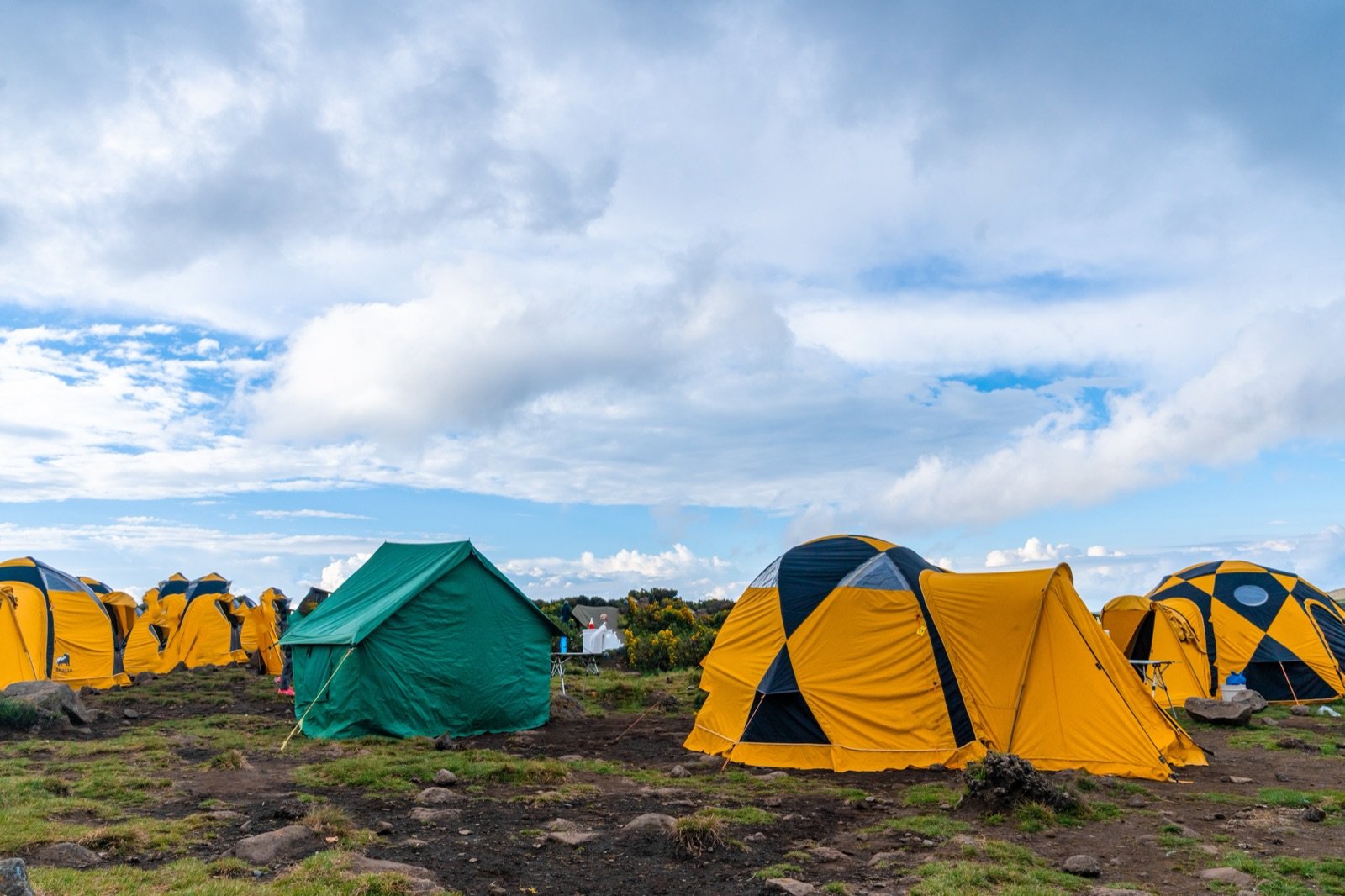 Yellow and green dome tents and a green canvas tent pitched on open rocky ground at a mountain campsite under a cloudy blue sky