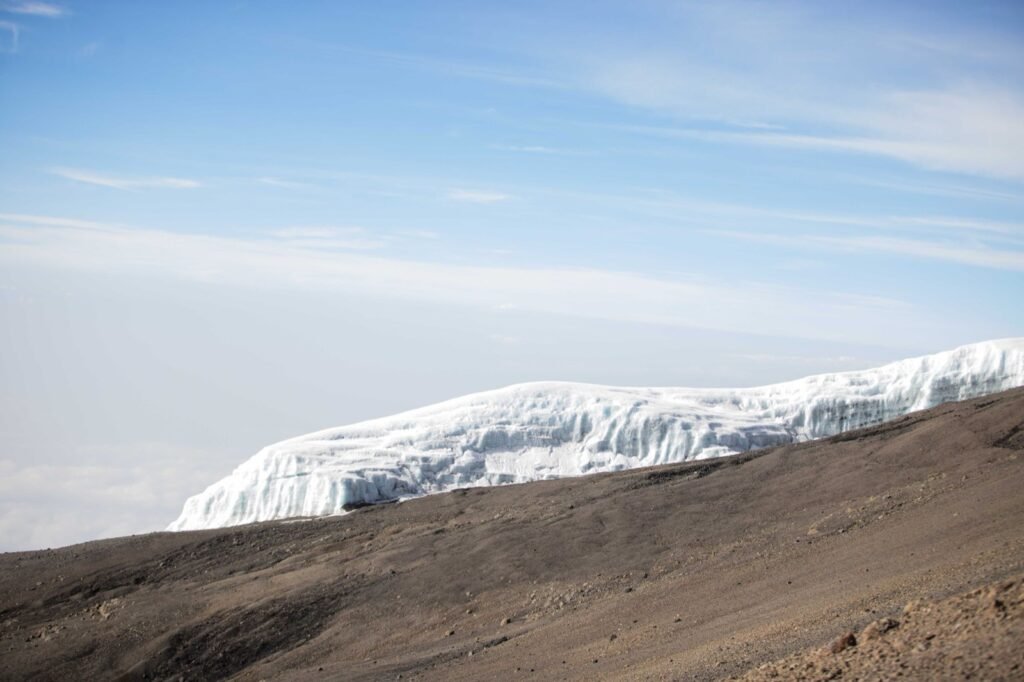 Wall of white glacier ice on the Kilimanjaro summit plateau above a slope of dark volcanic scree under a pale blue sky
