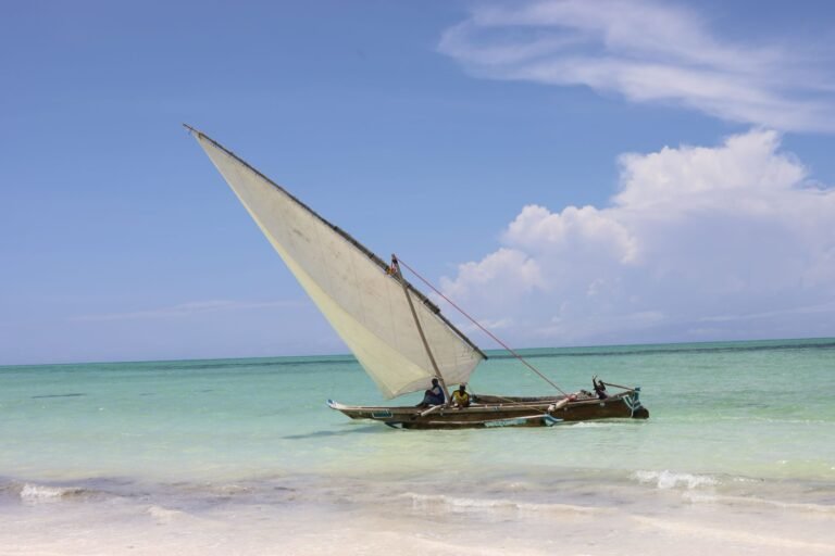 Traditional dhow with a white lateen sail and two people aboard on turquoise water just off a white-sand Zanzibar beach