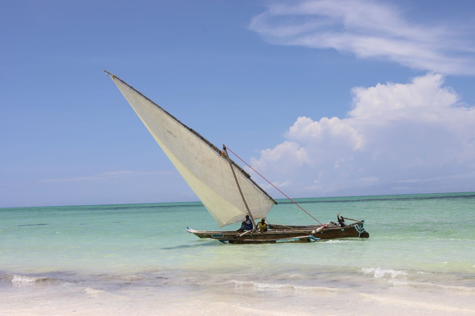 Traditional dhow with a white lateen sail and two people aboard on turquoise water just off a white-sand Zanzibar beach